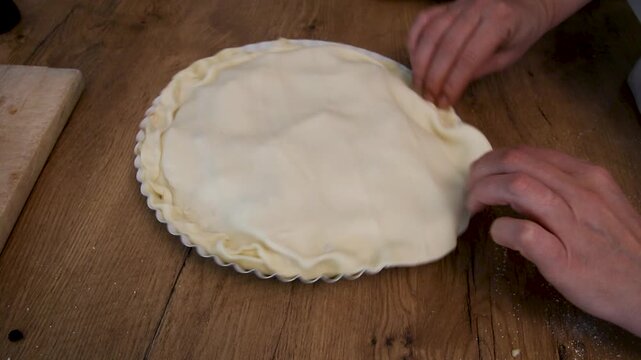 a person presses and shapes raw pie dough edges by hand on a wooden table in a home kitchen during baking preparation to seal and form a pie