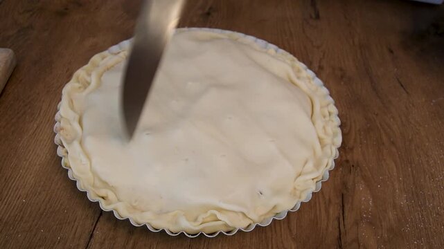 a person cuts raw pie dough with a knife in a home kitchen on a wooden table during food preparation to make a baked pie