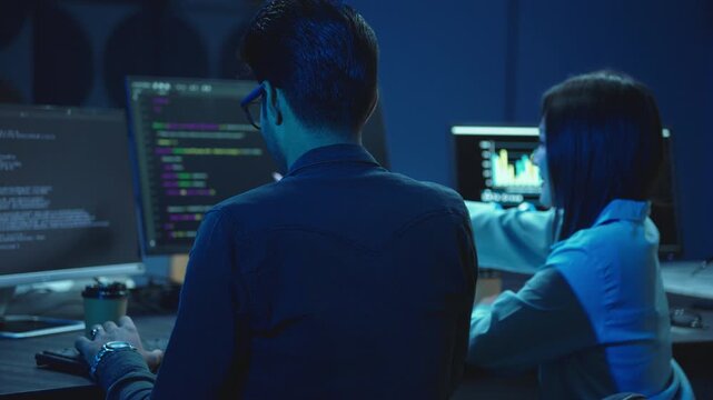 Back view of two developers at desks with code and data on monitors, woman pointing at screen while discussing project in a dark blue office at night.