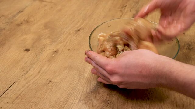 a person mixes raw chicken with marinade by hand in a bowl on a wooden table in a home kitchen during cooking preparation to evenly coat meat before baking
