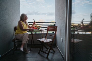 Obraz premium A mature woman sits on a small balcony holding a smartphone beside a plate of fresh fruit. She relaxes on wooden chairs while checking her phone and enjoying the view.