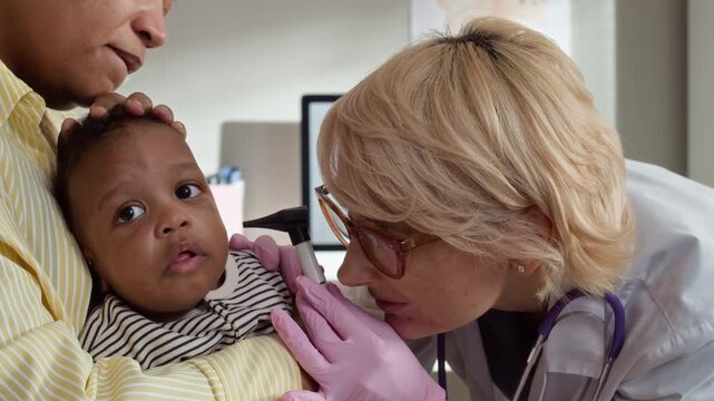 Medium closeup of Caucasian pediatrician examining African American baby held by African American mother using otoscope. Ideal for pediatric clinic ads and healthcare marketing campaigns