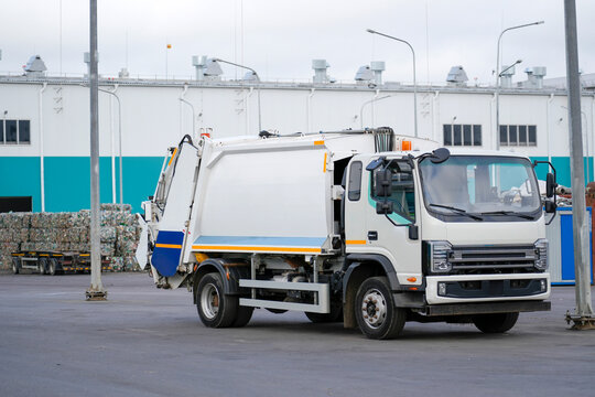 White garbage truck is parked, featuring its hydraulic compaction mechanism. Other trucks and industrial structures