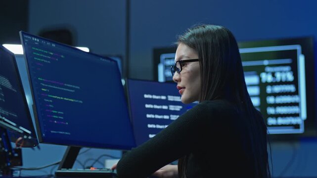 Asian woman with long hair working at desk with several screens showing program code and data statistics, software development in dark office.