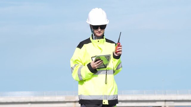 Engineer inspection for industrial safety, hardhat high visibility, radio and tablet checklist, bridge site walkie talkie technician recording maintenance compliance data under clear blue sky
