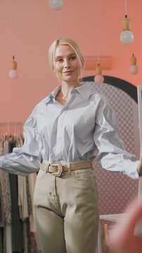 Stylish young blonde woman adjusting shirt collar while looking at her reflection in a mirror inside a modern fashion boutique. Female shopper is trying on an outfit and evaluating her look in a soft