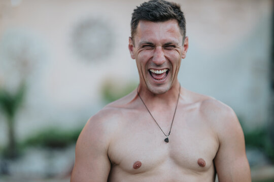Shirtless young man laughing with eyes closed showing a wide smile and yin yang tattoo. Casual outdoor portrait capturing joyful, candid emotion and relaxed energy.