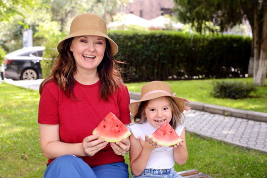 Happy mother and daughter holding watermelon slices in a sunny park. Healthy summer snack, family bonding, joyful childhood, outdoor lifestyle, and wellness concept