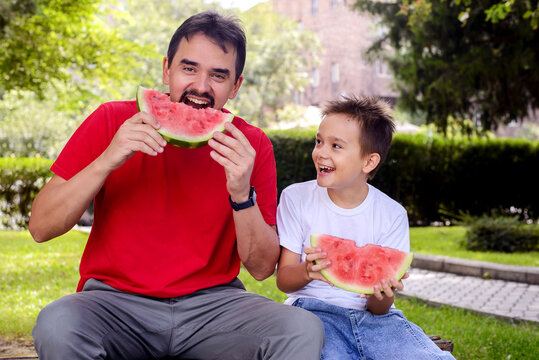 Happy father and son eating watermelon in a sunny park. Fun family bonding, healthy summer snack, joyful childhood memories, outdoor lifestyle, and wellness concept