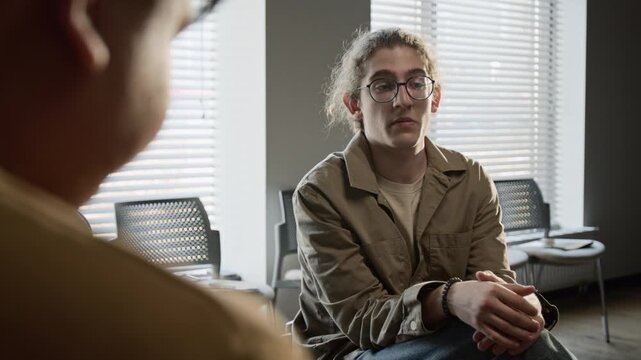 Over the shoulder portrait of teenager with glasses sitting in group therapy session and looking at camera. Footage for mental health awareness campaigns and youth counseling services