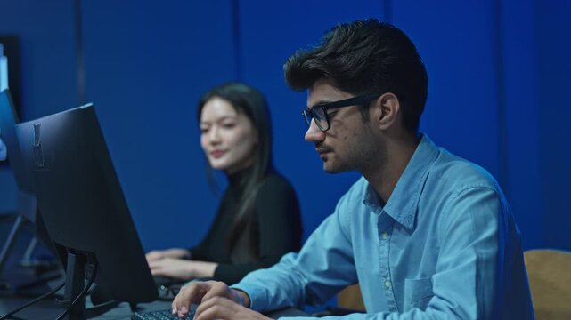 Young Indian man with glasses using computer in blue office, female coworker typing behind, international IT team working late at night.