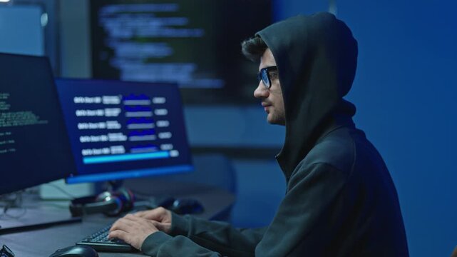 Young man in hooded sweatshirt and glasses typing on keyboard with data on monitors, hacking and cyber crime concept in dark room.