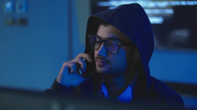 Young man in black hooded jacket with glasses making phone call in dark room with screens, cyber crime, fraud and phone scam concept.