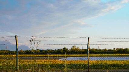 Fototapeta premium Wire mesh fence with barbed top overlooking a field and distant mountains