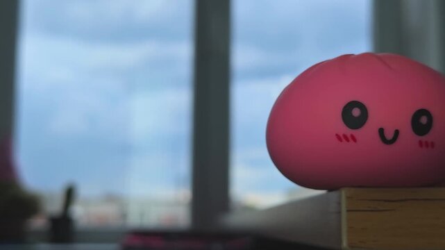 Pink squishy toy with a smiling face positioned on a wooden shelf, surrounded by soft lighting and a blurred background of a window and cityscape