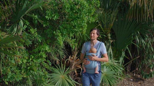 Father walking outdoors with infant son in ergonomic baby carrier. Slow motion.