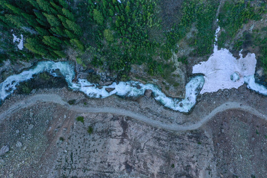 Aerial view of a rushing mountain river flowing alongside a winding dirt road through a dense evergreen forest with patches of snow CO, United States.