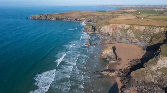 Aerial view of Bedruthan Steps with rugged cliffs, a sandy beach, and blue waves crashing against the rocky coastline near green fields in Padstow, Cornwall, United Kingdom.