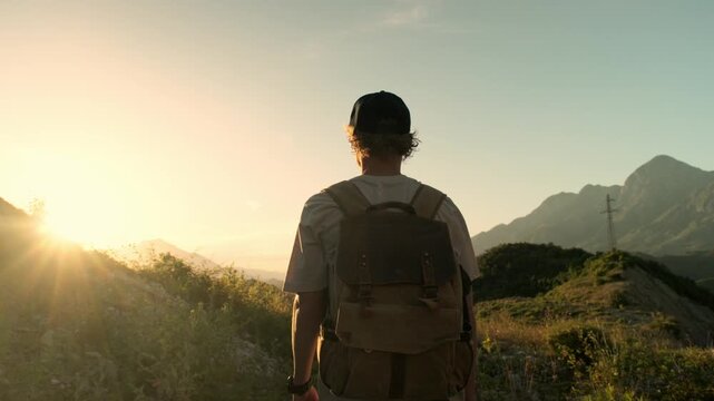 Handheld medium rear follow shot of male traveler with backpack walking to ridge and stopping to watch sunset mountains. Golden light and moving camera create immersive hiking mood outdoors.