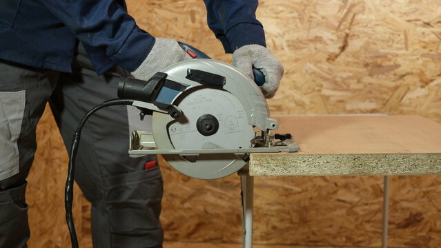 Close-up of worker hands cutting a wooden particle board with a handheld electric circular saw. Carpenter in work gloves operating power tools. Construction, carpentry workshop, and safety concept