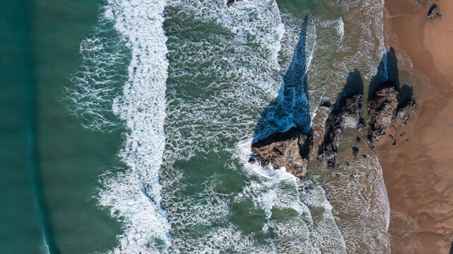 Aerial view of Bedruthan Steps with turquoise waves crashing against dark sea stacks and long shadows on the sandy beach in Padstow, Cornwall, United Kingdom.