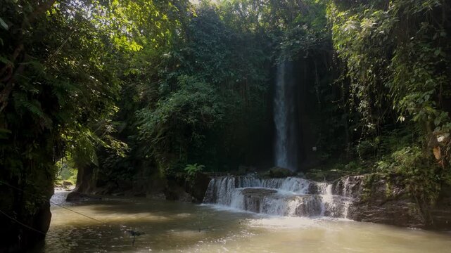 Sumampan Waterfall in the jungle of Bali, bathed in sunlight. Slow motion static shot. The waterfall flows into the river and valley.