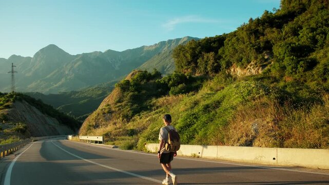 Handheld wide shot of male traveler walking downhill in the middle of mountain road at sunset. Distant figure, winding asphalt and vast peaks emphasize freedom, journey and solitude outdoors.