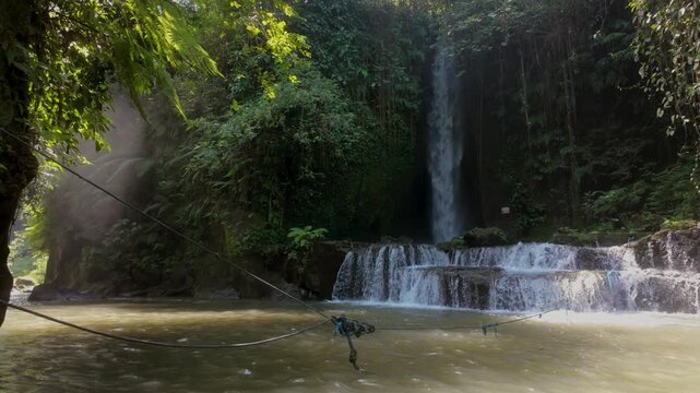 Sumampan Waterfall in the jungle of Bali, bathed in sunlight. Slow motion tracking forwards shot. The waterfall flows into the river and valley.