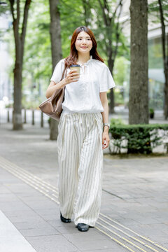 テイクアウトコーヒーを片手に緑の並木道を歩く日本人女性