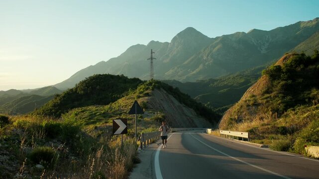 Handheld wide frontal shot of male traveler walking uphill along mountain road, then pausing to look around and catch breath. Golden light and expansive landscape emphasize effort and adventure.