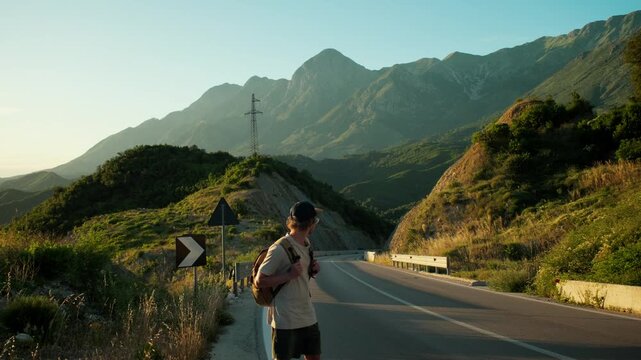 Handheld wide shot of male traveler standing on mountain road, scanning for cars and extending arm to hitchhike. Backpack, roadside curve and sunset peaks frame waiting and travel uncertainty.