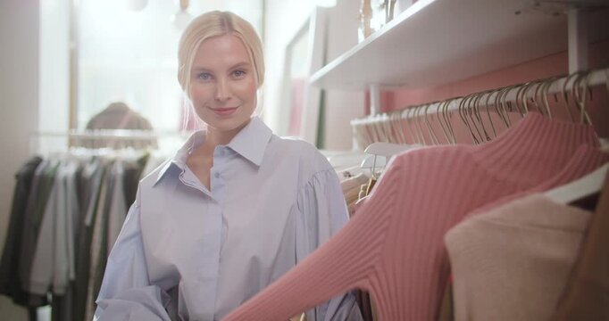Stylish young blonde woman choosing a pink sweater while browsing clothes in a modern fashion boutique. Female shopper examines knitwear on a clothing rack in a soft pastel retail interior with