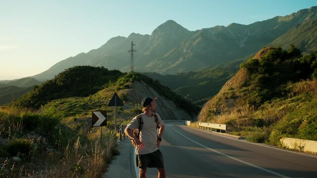 Handheld wide shot of male traveler waiting for ride on empty mountain road, then looking around and leaving frame. Still roadway, backpack and sunset peaks show failed hitchhiking attempt.