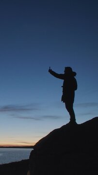 Silhouette of a man standing on a rocky cliff and taking a selfie with smartphone at twilight by a calm lake. Dark figure against blue evening sky and distant horizon creates a dramatic outdoor travel