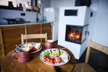 Obraz premium Cozy breakfast in rustic kitchen with fireplace, plates of eggs, avocado, prosciutto and coffee on wooden table, concept of hygge lifestyle, home comfort, warm morning routine