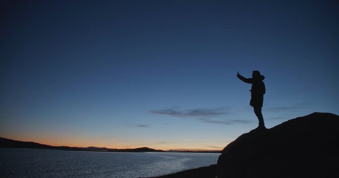 Silhouette of a man standing on a rocky shore and taking a selfie with a smartphone at twilight. Dark figure against a deep blue evening sky and calm water creates a dramatic outdoor travel scene with
