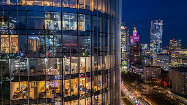 Aerial view of the illuminated glass facade of a modern skyscraper with office interiors and the Warsaw city skyline featuring the Palace of Culture and Science at night in Poland.
