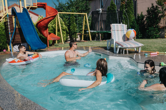 A group of friends relax and play in a backyard pool with inflatable floats. The summer scene shows casual fun by a slide and lounge chair in bright sunlight.