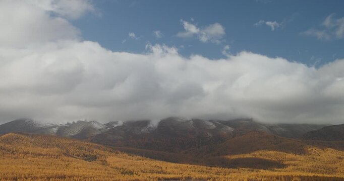 Timelapse of large clouds moving over autumn mountains with golden forest and snow patches on the peaks. Changing weather and shifting light create a dramatic natural landscape with seasonal contrast