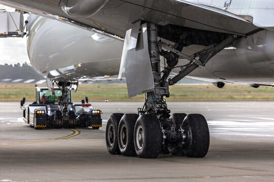 Widebody aircraft undergoes pushback operation wet airport apron heavy tow tractor maneuvers fuselage. Detail landing gear assembly rain aviation engineering ground support operational efficiency
