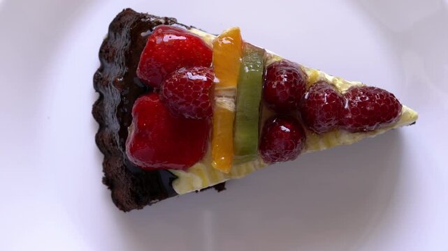Close-up of glossy fruit tart slice with berries, citrus and chocolate crust on white plate