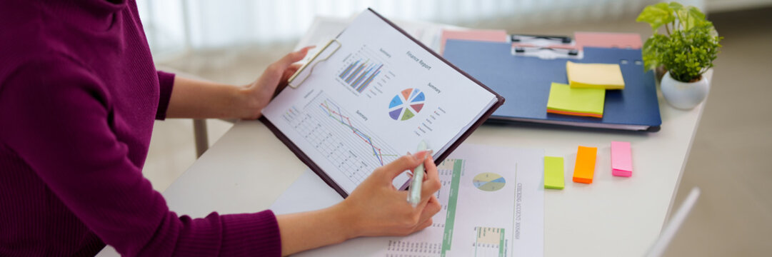 Young Asian professional analyzing colorful business charts and data reports at modern bright office workspace