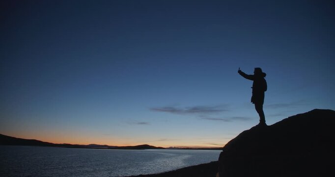 Silhouette of a man standing on a rocky cliff and taking a selfie with smartphone at twilight by a calm lake. Dark figure against blue evening sky and distant horizon creates a dramatic outdoor travel
