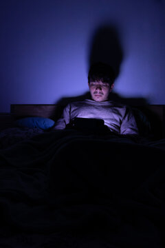 Relaxed man lying in bed using his cell phone in a dark room. The screen light illuminates his face in a nighttime scene of rest and digital connection