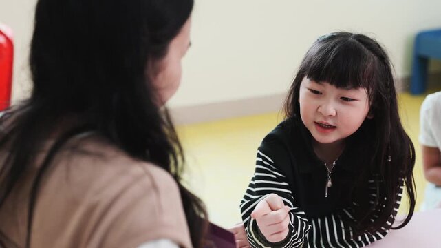 Smiling Asian girl listening to teacher or mother in kindergarten. Child communicating and learning with adult, showing attention, trust and care in early education classroom.