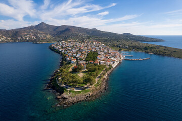 Fototapeta premium Drone shot of the traditional coastal village of Perdika on Aegina island. White houses and a scenic marina stretch along a rocky peninsula into the deep blue Saronic sea, with mountains in background