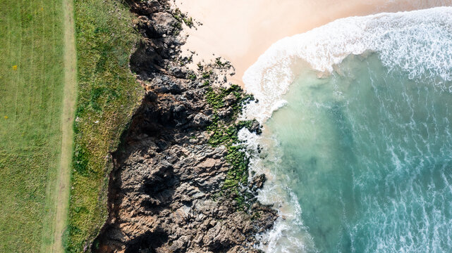 Aerial view of the rugged coastline at Trevone featuring grassy cliffs, dark mossy rocks, and turquoise waves crashing onto a sandy beach Padstow, Cornwall, United Kingdom.