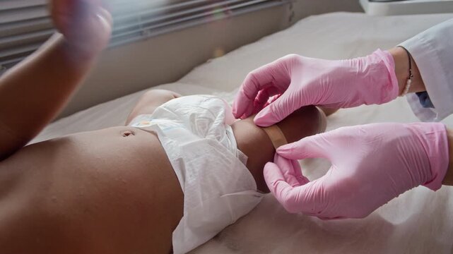 Closeup of pediatrician wearing pink gloves applying adhesive bandage on leg of infant lying on examination table. Footage is ideal for pediatric healthcare and vaccination campaign commercials