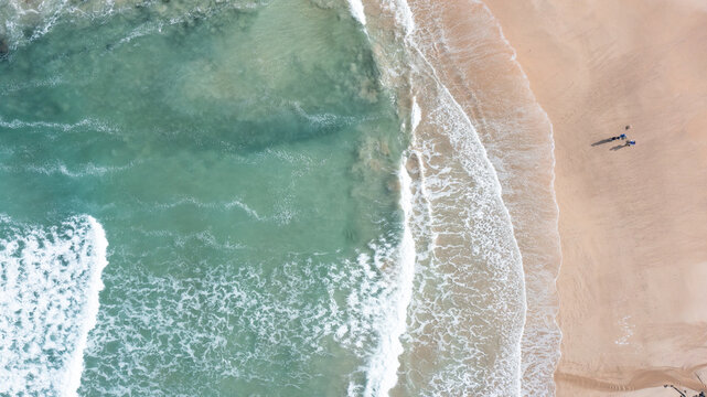 Aerial view of turquoise waves crashing on the golden sand of Trevone Bay with people walking on the beach in Padstow, Cornwall, United Kingdom.