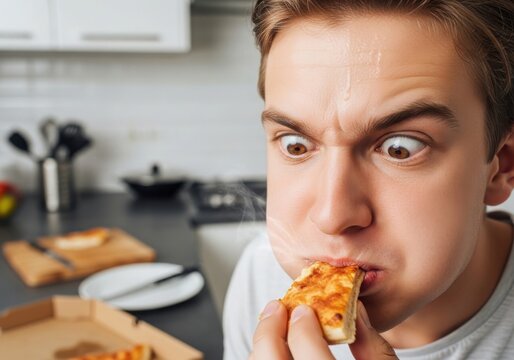 Closeup of a young man with a funny expression eating a hot slice of pizza in a kitchen